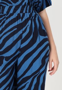 Zila un tumši zili zebra svītroti audumi ar elastīgu jostasvietu, kas izceļas ar gludu, mīkstu tekstūru un brīvu piegriezumu, daļēji redzama roka.