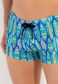 Blaue Schwimmshorts mit einem Blattmuster in Grün, Orange und Gelb. Sie verfügen über einen schwarzen Kordelzug und ein figurbetontes Design.