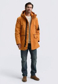 Orangefarbener Parka mit abnehmbarer Kapuze, Frontreißverschluss und mehreren Taschen. Kombiniert mit einem cremefarbenen Pullover und blauen Jeans.