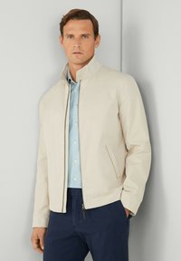 Beige jakke med høj krave, lynlås lukning og sidelommer, båret over en lyseblå skjorte med knapper og mørkeblå bukser.