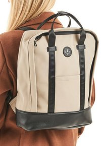 Beige rugzak met een textuur van zwart leer aan de onderkant, zwarte riemen en een ritssluiting. Voorzien van een voorvak en een rond logo.