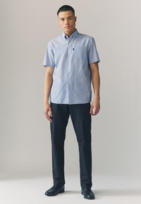 Kortærmet skjorte med knapper foran med blå og hvide striber, der har en brystlomme. Kombineret med mørke habitbukser og sorte sko.