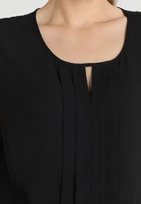 Blusa nera con scollatura rotonda e una piccola apertura a forma di chiave. Presenta pieghe verticali che scendono lungo la parte anteriore. Tessuto dalla superficie liscia.
