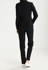 Czarny garnitur na miarę z dopasowaną marynarką i spodniami o prostych nogawkach. Gładka tkanina, czyste linie i minimalistyczny design. Noszony z białymi trampkami.