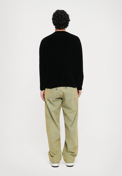 Man with curly dark hair wearing a black sweater, light green loose jeans, and white sneakers standing facing a white background.