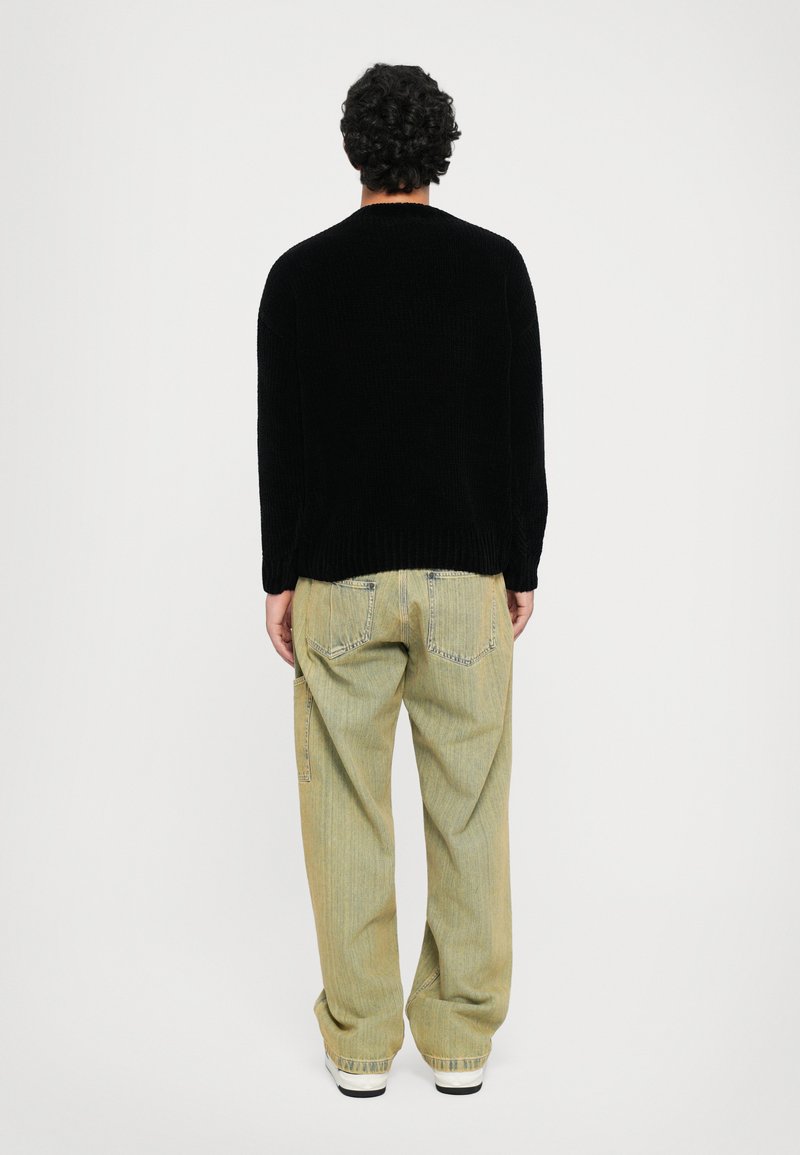Mand med krøllet mørkt hår iført en sort sweater, lysegrønne løse jeans og hvide sneakers, der står foran en hvid baggrund.