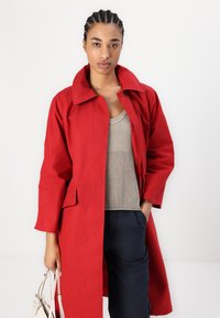 Czerwony płaszcz z długim rękawem, szerokim kołnierzem i przednimi kieszeniami, noszony na jasnoszarym dzianinowym topie i ciemnoniebieskich spodniach. Charakteryzuje się luźnym krojem.