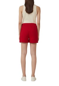 Pantaloni corti rossi in morbido tessuto con vita elastica, abbinati a una canotta di colore chiaro e scarpe da ginnastica bianche. Vista da dietro.