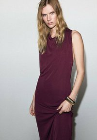 Robe bordeaux sans manches en tissu doux, avec un décolleté drapé et un léger effet froncé. Portée avec un bracelet jonc en or.