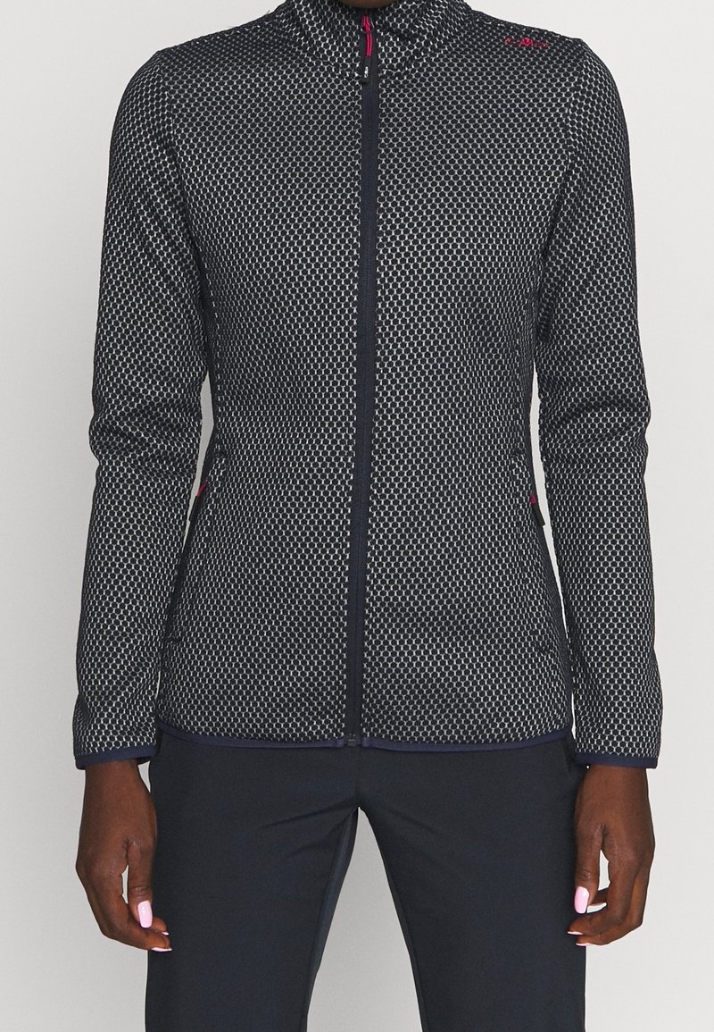 Personne portant une veste zippée à motif noir et blanc avec manches longues et un pantalon noir, montrée du cou aux hanches.