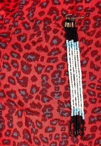 Rød stof med leopardmønster, prydet med en sikkerhedsnål og tråde af perler i sort, hvid og lysblå hængende fra en lomme.