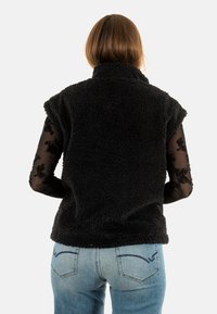 Zwarte, pluche, mouwloze fleecestas met een hoge kraag. Gecombineerd met een doorzichtige lange mouwen top. Draagt met lichtblauwe jeans met achterzakken.