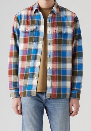 Mężczyzna ubrany w rozpiętą, wielokolorową koszulę w kratę na brązowym t-shircie i jasnoniebieskich dżinsach, stojący na jednolitym tle.