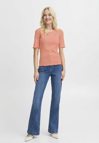 Koral ribbet strikbluse med flagrende ærmer, parret med flagrende blå denimjeans. Hvide hæle komplementerer ensemblet.