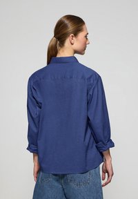 Mörkblå knapp-up skjorta med uppvikta ärmar, avslappnad passform och detalj på bakstycket, tillsammans med ljusblå högmidjade denimjeans.