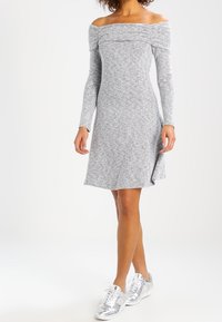 Donna che indossa un vestito grigio a spalle scoperte e maniche lunghe, abbinato a sneakers argentate brillanti, in piedi su uno sfondo bianco semplice.