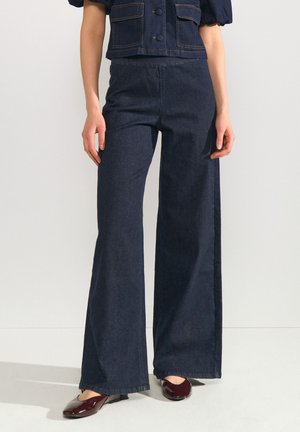 Donna che indossa jeans a vita alta blu scuro a gamba larga con scarpe piatte bordeaux e giacca corta blu scuro coordinata.