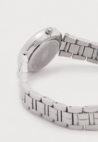 Ceas din metal argintiu cu o carcasă rotundă și coroană texturată. Brățara are zale dreptunghiulare, cu finisaje lustruite și satinate.