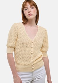 Cardigan en tricot jaune clair avec un col en V, des manches courtes bouffantes, des motifs texturés et des boutons recouverts de tissu sur le devant.