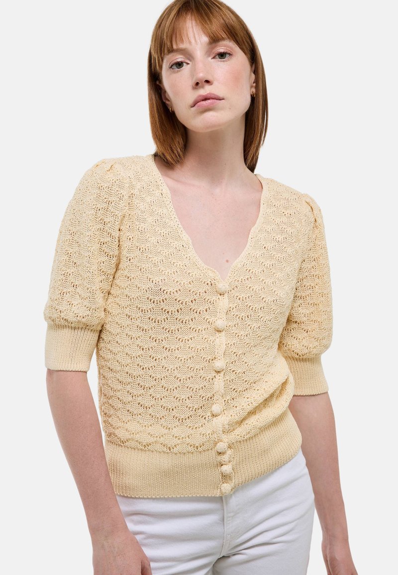 Cardigan en tricot jaune clair avec un col en V, des manches courtes bouffantes, des motifs texturés et des boutons recouverts de tissu sur le devant.