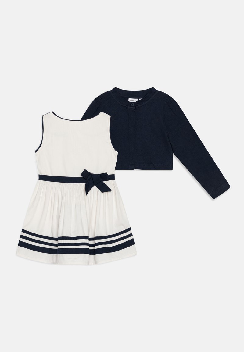 White dress with navy stripes, a bow at the waist, and sleeveless design; accompanied by a cropped navy cardigan with button closure.
