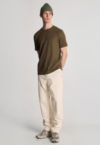 T-shirt verde oliva a maniche corte, pantaloni beige e sneakers grigio chiaro. Ambientato su uno sfondo bianco semplice. Design semplice e informale.