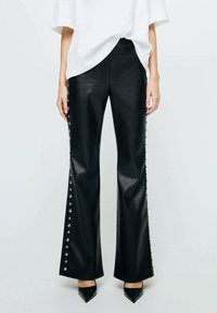 Pantalons évasés en simili cuir noir avec des accents de clous argentés le long des côtés. Texture lisse et design à jambes larges associés à des chaussures pointues noires.