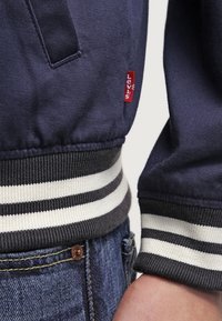 Veste bleu marine avec une ceinture côtelée grise et blanche à rayures. Présente une étiquette rouge Levi's et est fabriquée en tissu lisse.