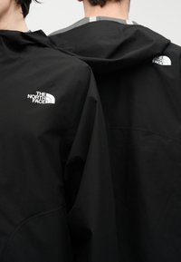 Δύο άτομα φορούν μαύρα μπουφάν The North Face με ορατά λογότυπα στον ώμο και την πλάτη, εμφανιζόμενα από πίσω και πλάγια.