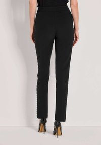 Zwarte fitted broek met een gladde textuur, voorzien van geschulpte details langs de zijkanten. Gecombineerd met hoge hakken.