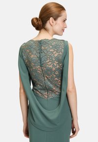 Grünes Kleid mit Spitzenrücken-Detail, das ein drapiertes Design und einen verdeckten Reißverschluss aufweist. Die glatte Stoffoberfläche ergänzt die filigrane Blumenspitze.