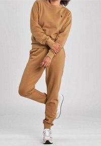 Brązowy dres z bawełny, składający się z bluzy i spodni dresowych z prążkowanymi mankietami, z małym logo na klatce piersiowej. Połączony z białymi i szarymi trampkami.
