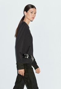 Zwarte lange mouwen top met een gevlochten leren riem op de taille. G gedragen met zwarte gestructureerde broek, die een strakke, minimalistische uitstraling toont.