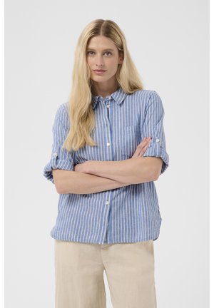 Blonde Frau mit langen Haaren, die ein blau-weiß gestreiftes Hemd mit hochgekrempelten Ärmeln und beige Hose trägt, steht mit verschränkten Armen.