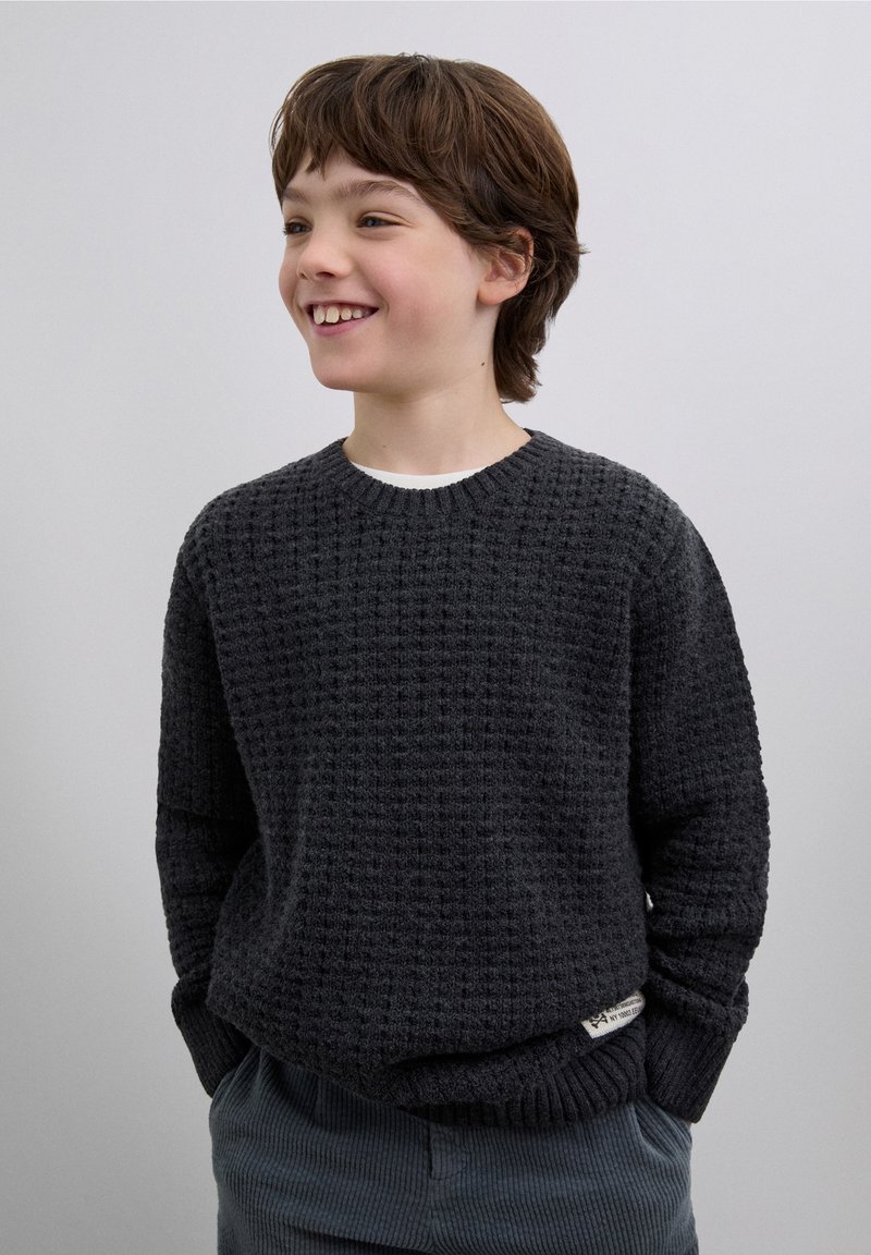 Pull en maille texturée gris foncé avec un col rond, doté de poignets et d'un ourlet côtelés, associé à un pantalon en velours côtelé bleu clair.