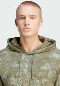 Huvtröja i olivgrön med en texturerad, splattrig mönster; har dragsnören och en minimalistisk logotyp på bröstet.