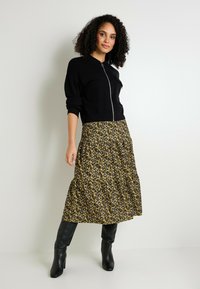 Sudadera negra con cremallera y dos bolsillos en el pecho, combinada con una falda larga amarilla con flores que presenta acentos en negro y botas de tacón alto negras.