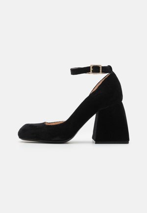 Melnas zamša zābaku kurpes ar papēdi un potītes siksniņu, kurai ir kvadrātveida metāla sprādze. Apaļš purngals un teksturēta virsma.