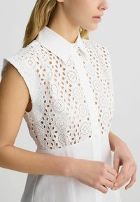 Blusa bianca con ricami, dotata di chiusura frontale con bottoni e colletto. Il tessuto presenta motivi ritagliati circolari e una texture liscia.