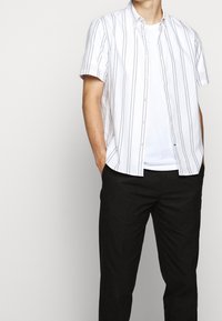 Muž stojící s rukama v kapsách, oblečený v bílém tričku, krátkém bílém sakě s tenkými černými pruhy a černých kalhotách.