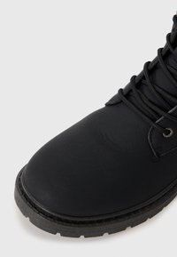 Botín de cuero negro con punta redondeada, acabado texturizado, ojetes de metal y cordones negros, que cuenta con una suela de goma resistente con patrones de tacos.