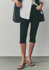 Leggings negros cortos con un diseño ajustado, combinados con tacones negros con tiras y una camisa blanca de botones, que muestran un look casual.