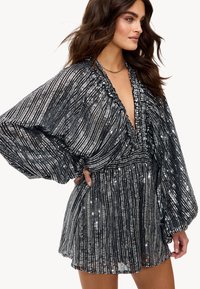 Robe en sequins noirs avec un décolleté en V profond et un col froncé. Manches longues et amples, taille cintrée, se terminant par une jupe plissée.