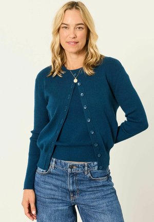 Blonde woman wearing a teal cardigan over a matching top and blue jeans, standing with one arm behind her back.