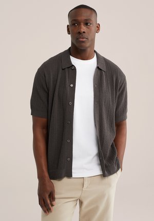 Homme portant une chemise tricotée à manches courtes gris foncé boutonnée sur un t-shirt blanc et un pantalon beige, debout avec une main dans la poche.