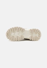 Suola di un sneakers beige e bianco con scanalature profonde e un logo impresso al centro su uno sfondo bianco.