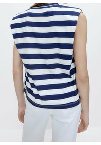 Una persona che indossa un top senza maniche a righe orizzontali blu navy e bianche, abbinato a pantaloni bianchi, vista di spalle su uno sfondo semplice.