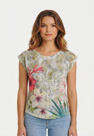 Donna con capelli lunghi e mossi indossa una maglietta a maniche corte con stampa floreale di foglie verdi e rosa e jeans azzurri, in piedi davanti a uno sfondo bianco.