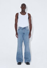 Jeans acampanados de mezclilla azul claro con cintura alta, combinados con una camiseta sin mangas blanca. Calzado negro y collares simples en capas.