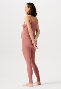 Schwangere Frau in einem rosa, figurbetonten Tanktop und Leggings steht barfuß, lächelt und hat die Hände hinter dem Rücken verschränkt vor einem weißen Hintergrund.